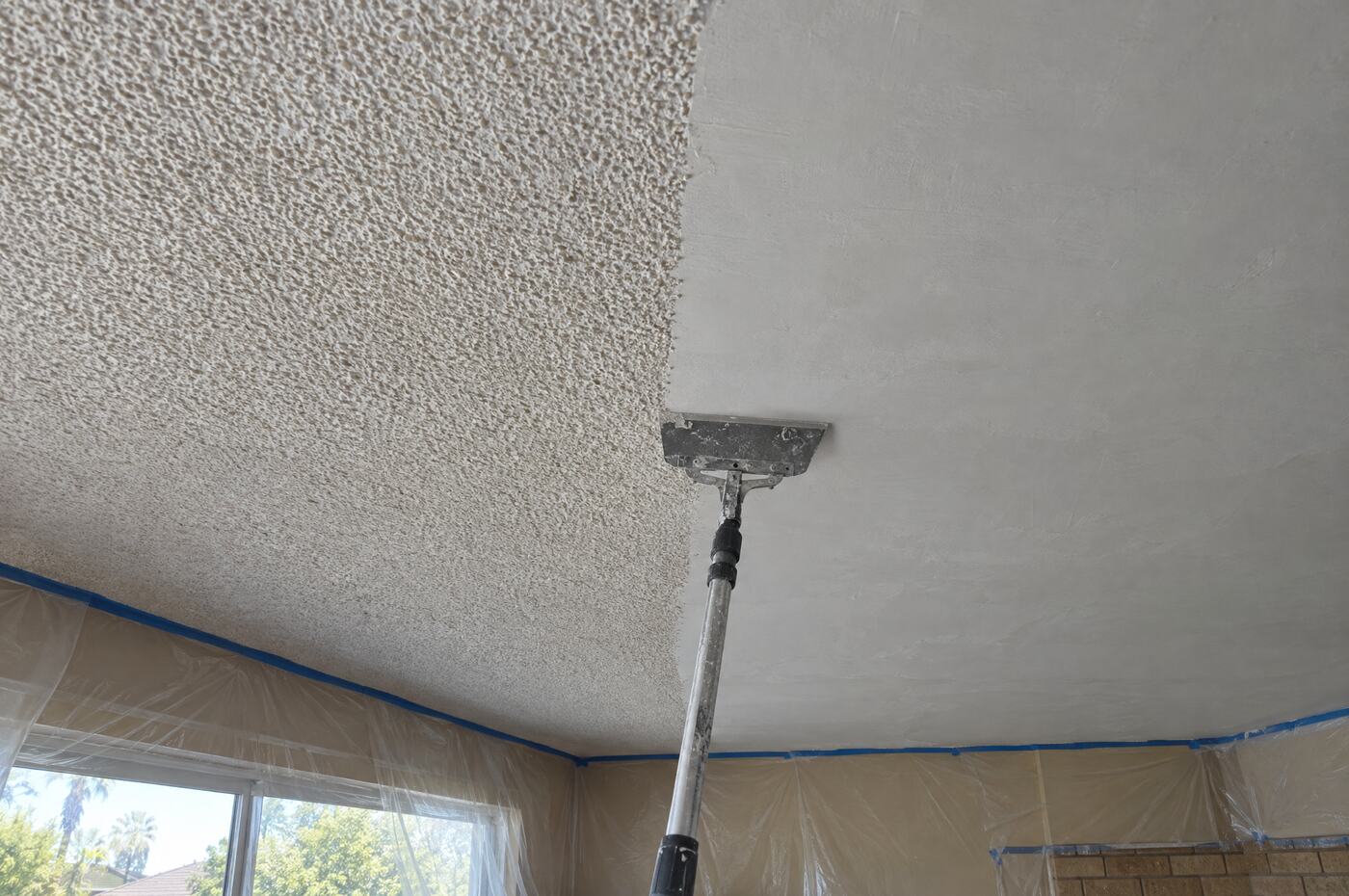 A ceiling halfway through popcorn removal: bumpy acoustic texture on the left, scraped smooth on the right, with a scraper on a pole at the line