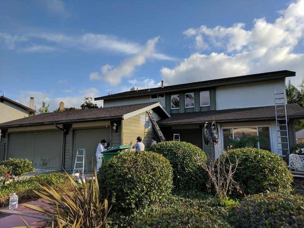 Two-story house with green-gray siding, ladders set up and painters prepping the garage and front